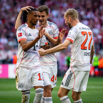 Tre giocatori del Bayern Monaco festeggiano dopo un gol. Sono sorridenti e si abbracciano, mostrando entusiasmo. Indossano maglie bianco e rosse con il logo della squadra e i numeri sul retro. Sullo sfondo si intravedono tifosi e un'atmosfera festosa.