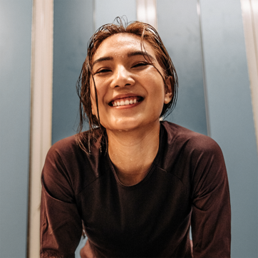 Una giovane donna sorridente con i capelli bagnati, indossa una maglietta scura. Si trova di fronte a uno sfondo blu chiaro, trasmettendo un senso di gioia e freschezza. La sua espressione riflette felicità e soddisfazione.