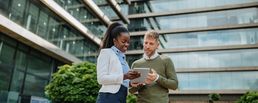 Two professionals engage in discussion outdoors. A woman in a white blazer and blue shirt looks at a tablet in her hands, while a man in a green sweater points at the screen, both smiling. The background features modern glass architecture and greenery.