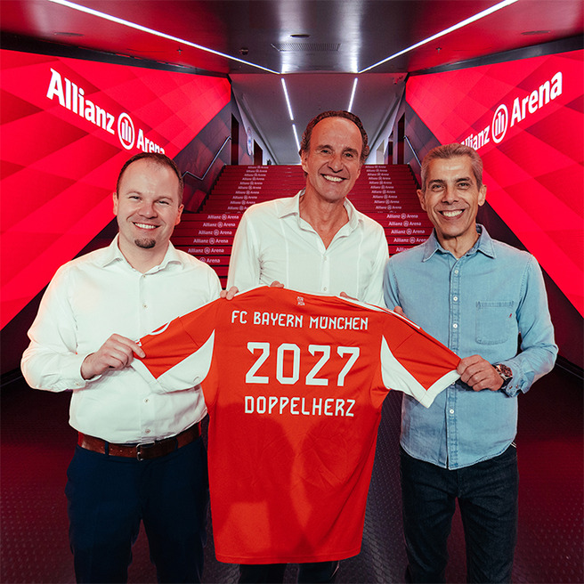 Tre uomini sorridenti sono in piedi all'interno dell'Allianz Arena, ognuno tiene in mano una maglia rossa del FC Bayern München. La maglia mostra il numero "2027" e la scritta "DOPPELHERZ". Sullo sfondo si vedono le scale illuminate con il logo dell'arena.