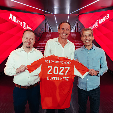Tre uomini sorridenti sono in piedi all'interno dell'Allianz Arena, ognuno tiene in mano una maglia rossa del FC Bayern München. La maglia mostra il numero "2027" e la scritta "DOPPELHERZ". Sullo sfondo si vedono le scale illuminate con il logo dell'arena.