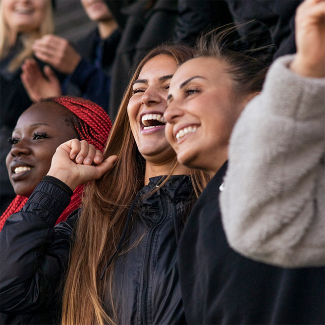 Tre giovani donne sorridenti si divertono insieme, esprimendo gioia e entusiasmo. Indossano abbigliamento casual scuro e uno dei volti è abbellito da trecce rosse. Il loro atteggiamento suggerisce un momento festoso e di condivisione.