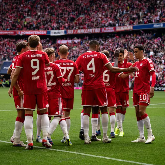 Giocatori del FC Bayern Monaco si riuniscono in un momento di celebrazione sul campo, indossando magliette rosse con numeri. La folla sullo sfondo è visibilmente entusiasta, creando un'atmosfera di festa dopo un'azione di squadra.